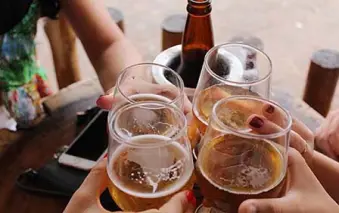 Four people clinking glasses of beer with a bottle in the center on a wooden table.